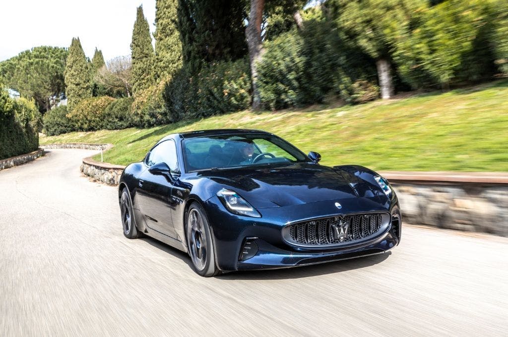 A brand new Maserati electric vehicle driving around a bend in the road in a leafy green area
