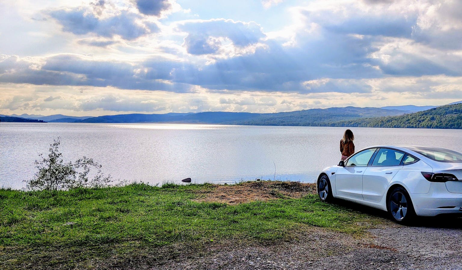 image shows woman standing next to electric car, next to lake with hills and clouds in the background